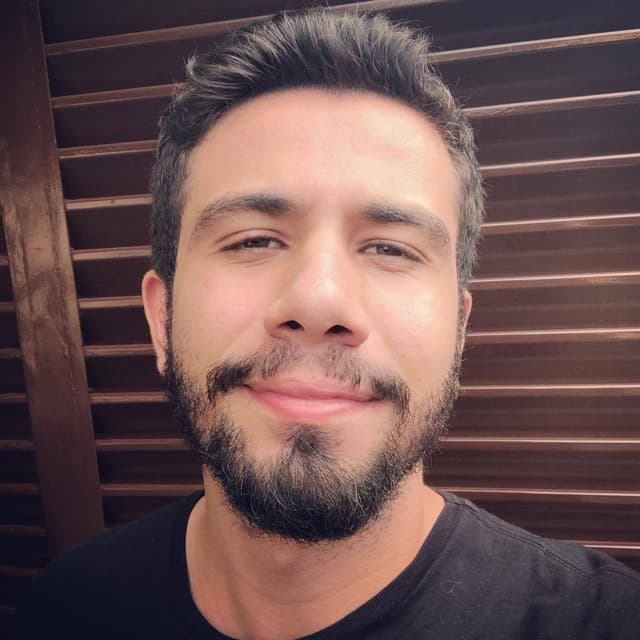 Selfie de Eduardo Paiva, homem branco com uma barba média, atrás da foto um portão eletrônico marrom